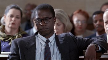 Movie still from “Primal Fear” (1996), directed by Gregory Hoblit – A man in a suit and tie sitting in front of a group of people; Close Up shot, Over the shoulder angle