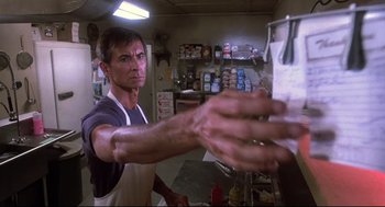 Movie still from “Psycho II” (1983), directed by Richard Franklin – A man in an apron reaches out to grab something from a counter; Medium shot, Low angle