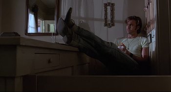 Movie still from “Psycho III” (1986), directed by Anthony Perkins – A man sitting on the edge of a dresser holding a wii controller; Wide shot, Low angle