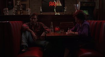 Movie still from “Psycho III” (1986), directed by Anthony Perkins – A man and a woman sitting at a table in front of a neon sign; Medium shot, Low angle
