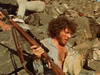Movie still from “Punishment Park” (1971), directed by Peter Watkins – A man holding a rifle while sitting on a rock pile; Medium shot, High angle