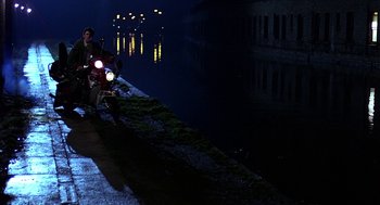 Movie still from “Quadrophenia” (1979), directed by Franc Roddam – A couple of people on a motorcycle at night by the water; Wide shot, High angle