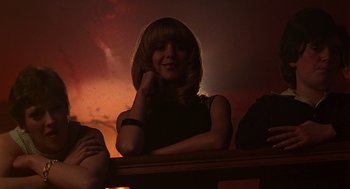 Movie still from “Quadrophenia” (1979), directed by Franc Roddam – A woman sitting on top of a railing in front of a sunset; Medium shot, Low angle