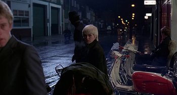Movie still from “Quadrophenia” (1979), directed by Franc Roddam – A woman standing next to a motorcycle on a street; Medium shot, Over the shoulder angle