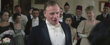Movie still from “Queen of the Desert” (2015), directed by Werner Herzog – A man in a tuxedo standing in front of a group of people; Medium shot, Low angle