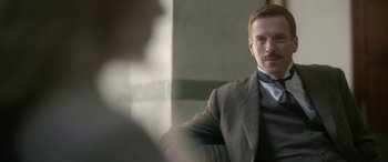 Movie still from “Queen of the Desert” (2015), directed by Werner Herzog – A man wearing a suit and tie sitting in front of a wall; Close Up shot, Over the shoulder angle