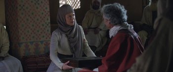 Movie still from “Queen of the Desert” (2015), directed by Werner Herzog – A woman wearing a headscarf is holding a box while two men watch; Medium shot, Over the shoulder angle
