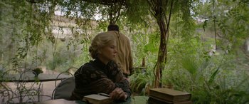 Movie still from “Queen of the Desert” (2015), directed by Werner Herzog – An older woman sitting at a table in a forest; Medium shot, Over the shoulder angle