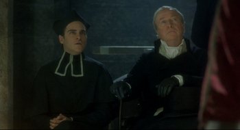 Movie still from “Quills” (2000), directed by Philip Kaufman – Two men sitting next to each other in black clothes; Medium shot, Low angle