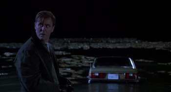 Movie still from “Raising Cain” (1992), directed by Brian De Palma – A man standing in front of a car in a parking lot at night; Medium shot, Over the shoulder angle
