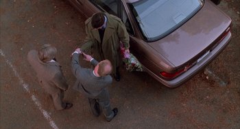 Movie still from “Raising Cain” (1992), directed by Brian De Palma – A man in a gray suit is handing flowers to another man in a brown coat; Wide shot, High angle
