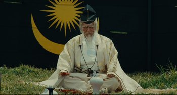 Movie still from “Ran” (1985), directed by Akira Kurosawa – An old man sitting on the grass with a white hat on; Medium shot, Low angle