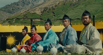 Movie still from “Ran” (1985), directed by Akira Kurosawa – A group of men sitting on the grass wearing hats; Wide shot, Low angle