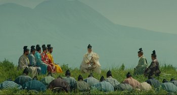 Movie still from “Ran” (1985), directed by Akira Kurosawa – A group of people sitting in a field with a mountain in the background; Extreme Wide shot, High angle