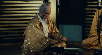 Movie still from “Ran” (1985), directed by Akira Kurosawa – An old man sitting on the ground wearing a robe; Medium shot, Low angle