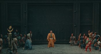 Movie still from “Ran” (1985), directed by Akira Kurosawa – A group of people sitting on the ground in front of a building; Extreme Wide shot, High angle