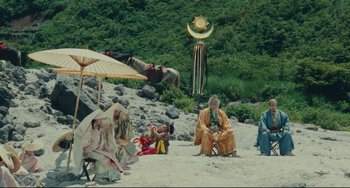 Movie still from “Ran” (1985), directed by Akira Kurosawa – A group of people sitting on a beach under an umbrella; Extreme Wide shot, Low angle
