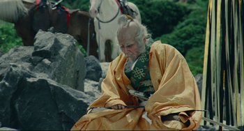 Movie still from “Ran” (1985), directed by Akira Kurosawa – An old man sitting on a rock near some horses; Medium shot, Low angle