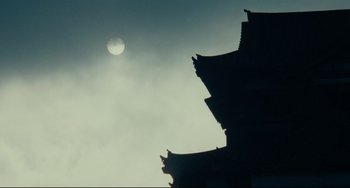 Movie still from “Ran” (1985), directed by Akira Kurosawa – The sun is setting in the sky behind a building; Extreme Wide shot, Low angle