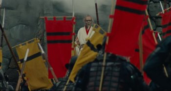 Movie still from “Ran” (1985), directed by Akira Kurosawa – An old man wearing a white robe standing in front of red and yellow flags; Wide shot, Over the shoulder angle