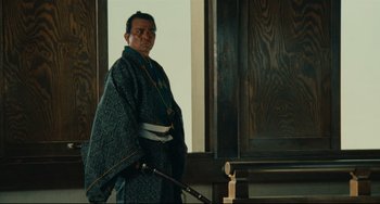 Movie still from “Ran” (1985), directed by Akira Kurosawa – A man standing in front of a wooden wall holding a sword; Medium shot, Low angle