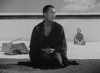 Movie still from “Rashomon” (1950), directed by Akira Kurosawa – A black and white photo of a man sitting on the ground; Wide shot, High angle