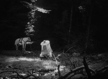 Movie still from “Rashomon” (1950), directed by Akira Kurosawa – A person in a white hat sitting in the grass next to a horse; Wide shot, Low angle