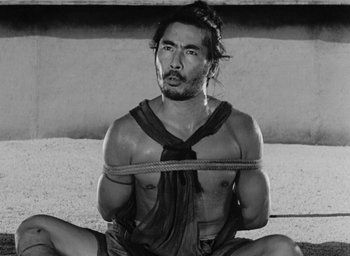 Movie still from “Rashomon” (1950), directed by Akira Kurosawa – A black and white photo of a man tied to a rope; Medium shot, High angle