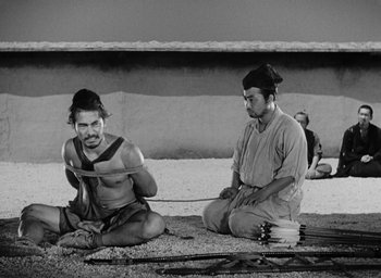 Movie still from “Rashomon” (1950), directed by Akira Kurosawa – Two men sitting on the ground in front of a wall; Wide shot, High angle