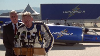 Movie still from “Rat Race” (2001), directed by Jerry Zucker – A man standing at a podium in front of an airplane; Medium shot, Low angle