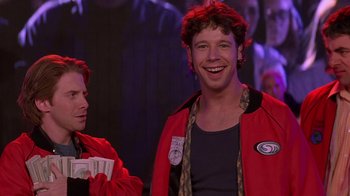 Movie still from “Rat Race” (2001), directed by Jerry Zucker – A man in a red jacket smiles at the camera while another man looks on; Medium shot, Over the shoulder angle