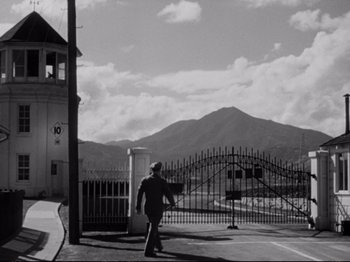 Movie still from “Raw Deal” (1948), directed by Anthony Mann – A man walking down a street near a fence; Extreme Wide shot, Low angle