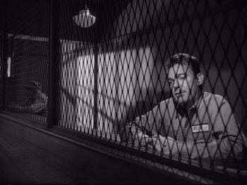 Movie still from “Raw Deal” (1948), directed by Anthony Mann – A black and white photo of a man behind a fence; Medium shot, Over the shoulder angle