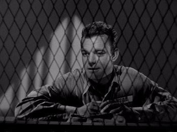 Movie still from “Raw Deal” (1948), directed by Anthony Mann – A black and white photo of a man behind a chain link fence; Medium shot, Low angle