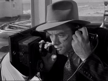 Movie still from “Raw Deal” (1948), directed by Anthony Mann – A black and white photo of a man in a hat and tie holding a camera; Close Up shot, Low angle