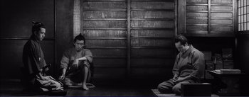 Movie still from “Red Beard” (1965), directed by Akira Kurosawa – A man sitting on the ground next to another man in front of a wooden wall; Wide shot, High angle