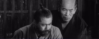 Movie still from “Red Beard” (1965), directed by Akira Kurosawa – A man sitting next to another man in a black and white photo; Close Up shot, High angle