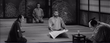 Movie still from “Red Beard” (1965), directed by Akira Kurosawa – A man sitting on the ground holding a sheet of paper in front of another man; Wide shot, High angle