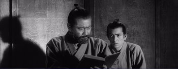 Movie still from “Red Beard” (1965), directed by Akira Kurosawa – A man reading a book while another man looks on; Medium shot, Low angle