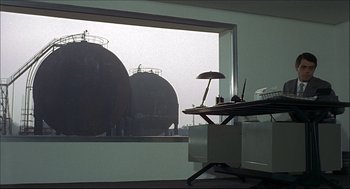 Movie still from “Red Desert” (1964), directed by Michelangelo Antonioni – A table with a lamp and a lamp on it in front of a window with a view of two oil tanks; Extreme Wide shot, Over the shoulder angle