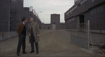 Movie still from “Red Desert” (1964), directed by Michelangelo Antonioni – Two men standing in an empty lot next to a building; Wide shot, Over the shoulder angle