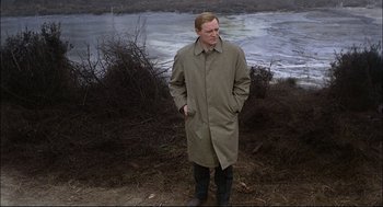 Movie still from “Red Desert” (1964), directed by Michelangelo Antonioni – A man in a trench coat standing in front of a body of water; Medium shot, High angle