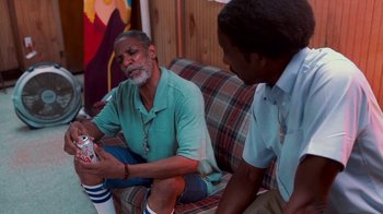 Movie still from “Red Hook Summer” (2012), directed by Spike Lee – Two men sitting on a couch and one of the men is holding a can of soda; Medium shot, Over the shoulder angle