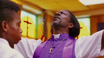 Movie still from “Red Hook Summer” (2012), directed by Spike Lee – A man in a priest's outfit with his arms outstretched in front of a stained glass window; Medium shot, Low angle