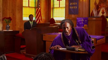 Movie still from “Red Hook Summer” (2012), directed by Spike Lee – An old man in a purple robe is sitting at a desk; Medium shot, Low angle