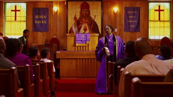 Movie still from “Red Hook Summer” (2012), directed by Spike Lee – A man in a purple robe stands in front of a painting of jesus; Wide shot, Low angle