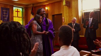 Movie still from “Red Hook Summer” (2012), directed by Spike Lee – A man and a woman hug in front of a group of people; Medium shot, Over the shoulder angle