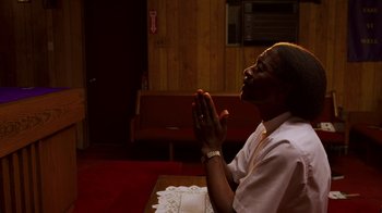 Movie still from “Red Hook Summer” (2012), directed by Spike Lee – A man sitting at a table with his hands in prayer; Medium shot, Low angle