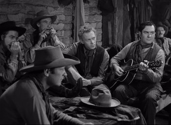 Movie still from “Red River” (1948), directed by Howard Hawks – A group of men sitting around a table playing guitars; Medium shot, Over the shoulder angle