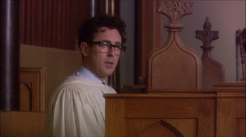 Movie still from “Reefer Madness: The Movie Musical” (2005), directed by Andy Fickman – A man wearing a robe and glasses in front of a podium in a courtroom; Medium shot, Low angle